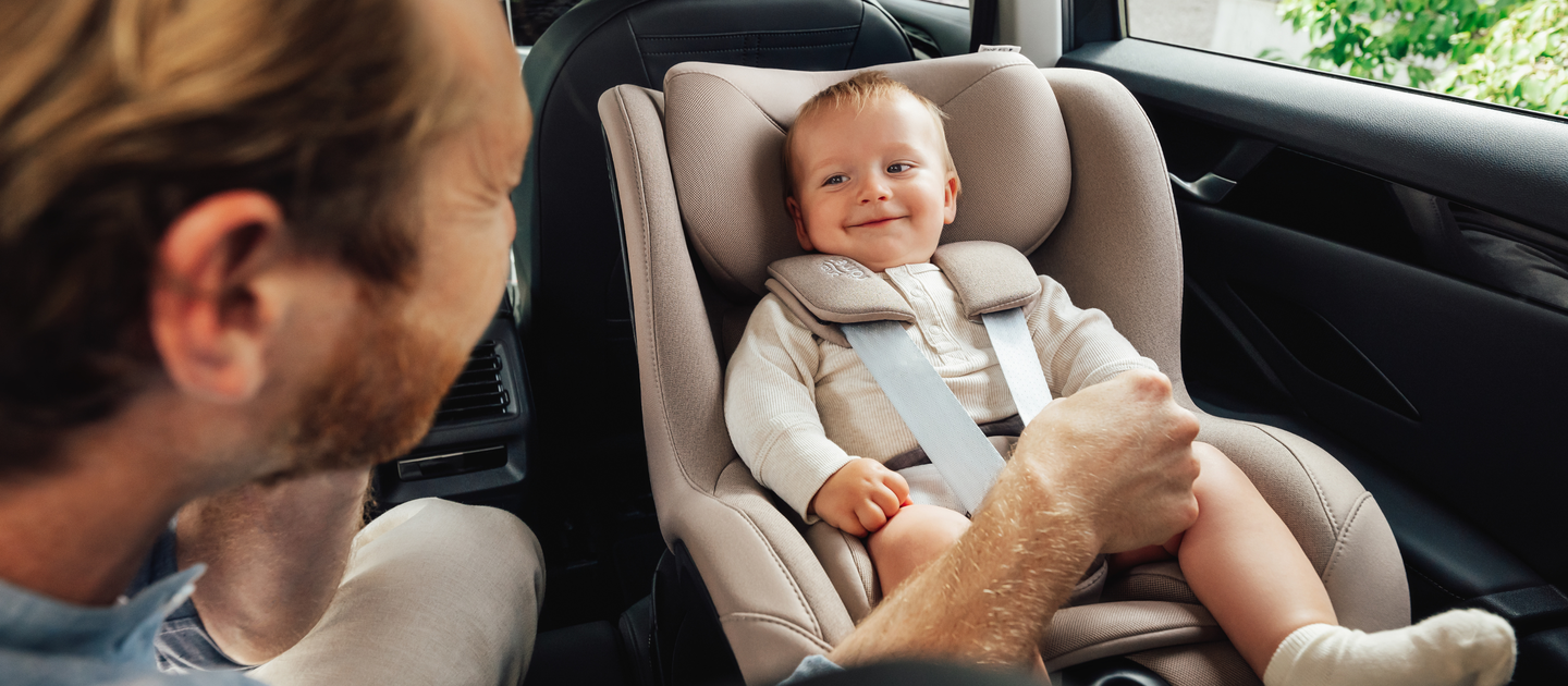 Um beb&ecirc; sorridente est&aacute; sentado com seguran&ccedil;a em uma cadeira automotiva Britax R&ouml;mer Dualfix 5Z na cor Teak, instalada no banco traseiro de um carro. Um adulto, visto de lado, segura suavemente a perna do beb&ecirc; enquanto interage com ele.